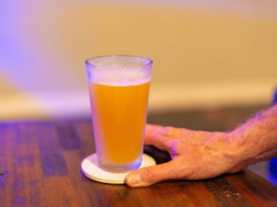 A male reaches for a tall clear glass of sour craft beer in a microbrewery. The cold refreshment has frost on the glass. The beer is on a table and coaster. The top of the glass has white froth.